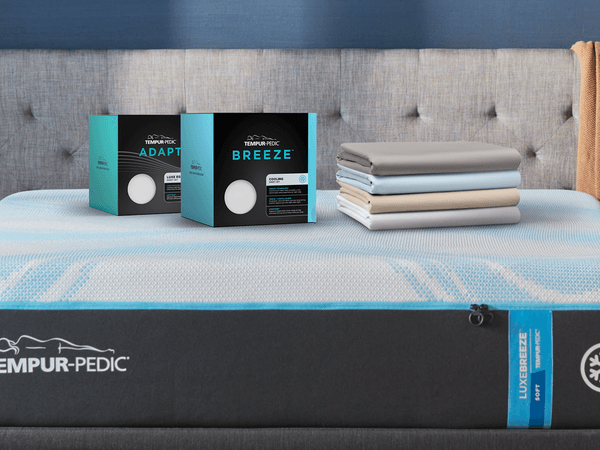 A Closeup of a TEMPUR-Breeze and TEMPUR-Adapt Sheet Set in a Box with a Stack of Folded Sheets Next to Them on a TEMPUR-LuxeBreeze Mattress in a Styled Room